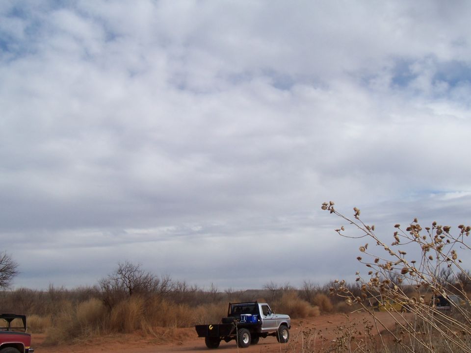 Silver flatbed truck driving on a dirt road with a red truck partially visible on the left