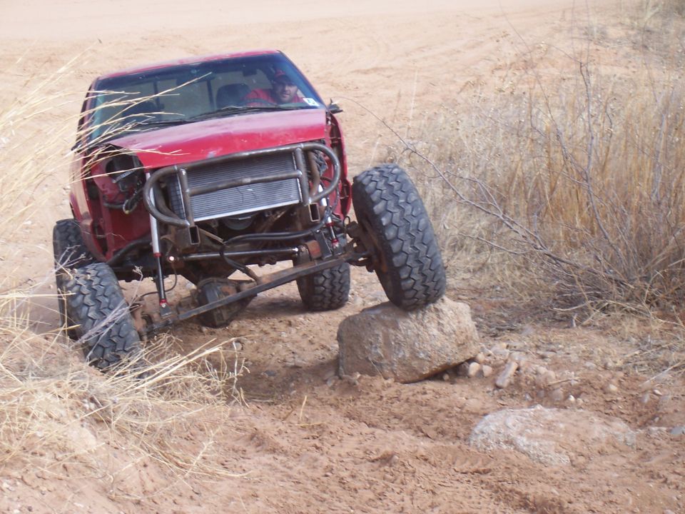 Red off-road truck with a custom front bumper and exposed radiator, one wheel on a rock