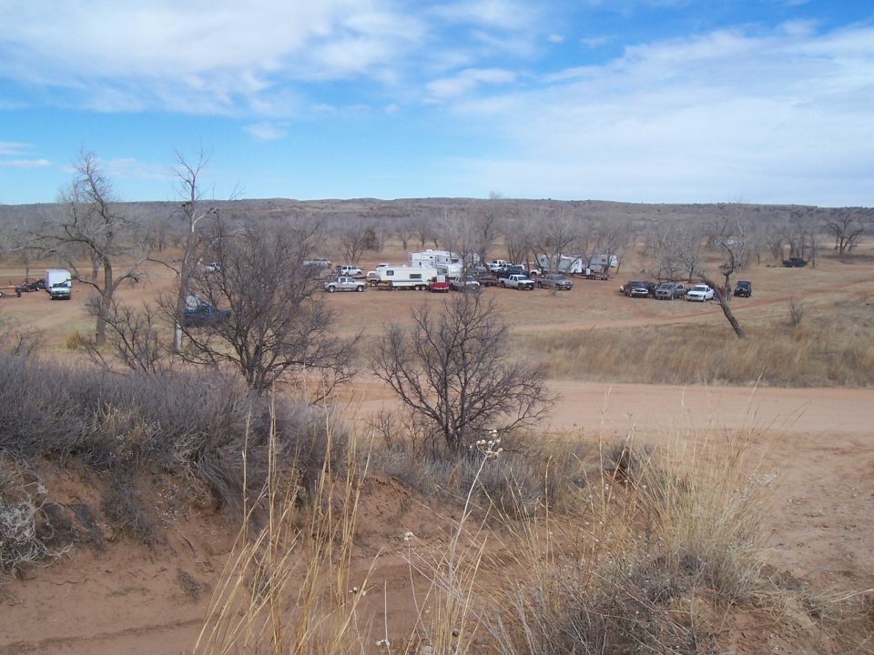 Dry, open field with numerous vehicles and RVs parked, bare trees, and a dirt road under a cloudy sky
