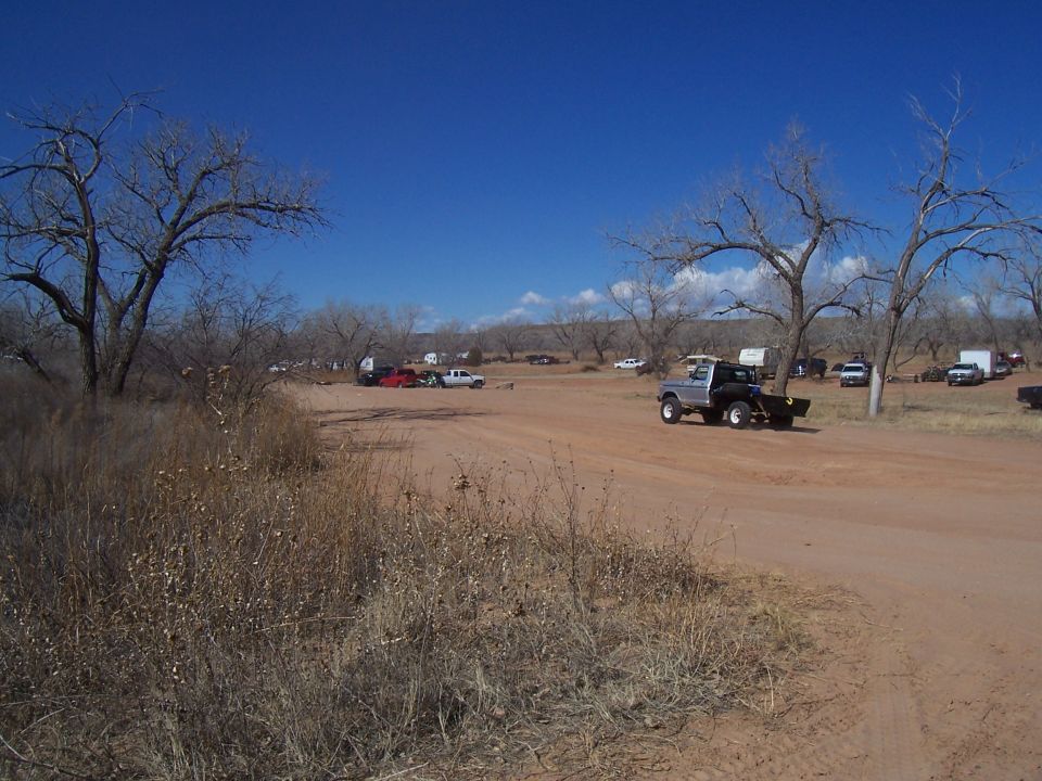 Several trucks and trailers parked in a dirt lot with bare trees and a bright blue sky