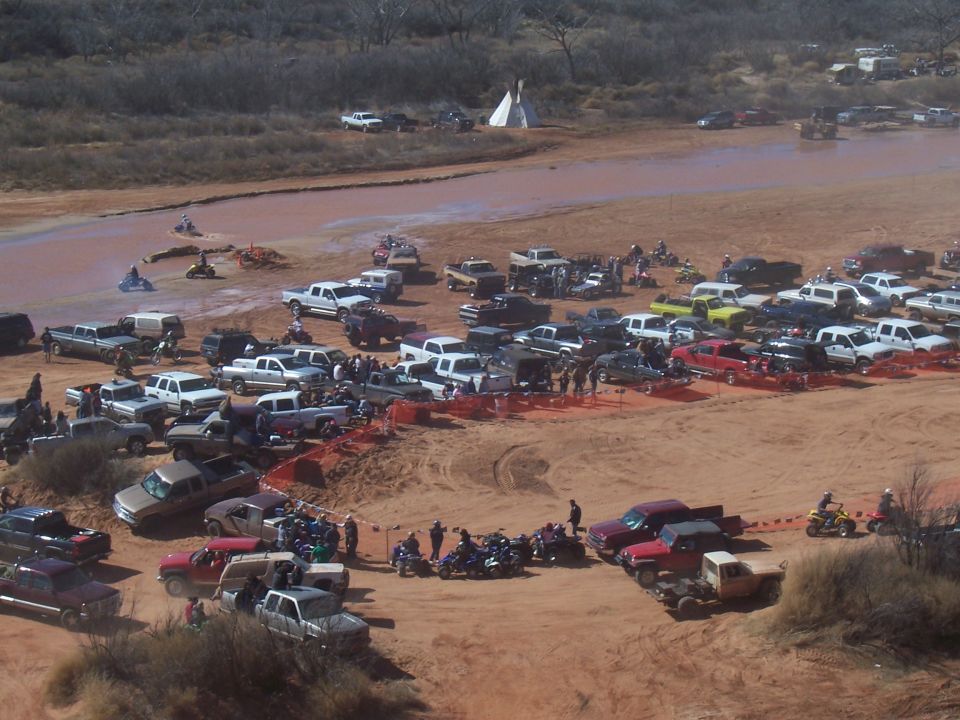 Dirt road scene with numerous trucks, ATVs, and motorcycles parked and driving in a muddy area