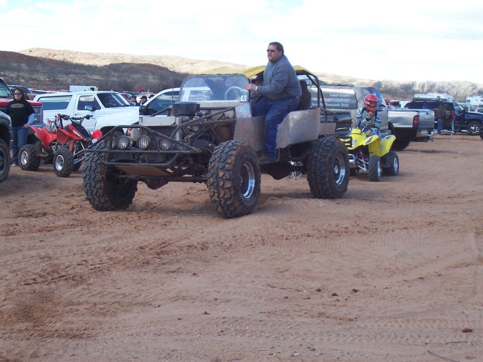 Custom off-road buggy with large tires, driven by a man, with a yellow ATV carrying a child behind it. Other vehicles and people are in the background