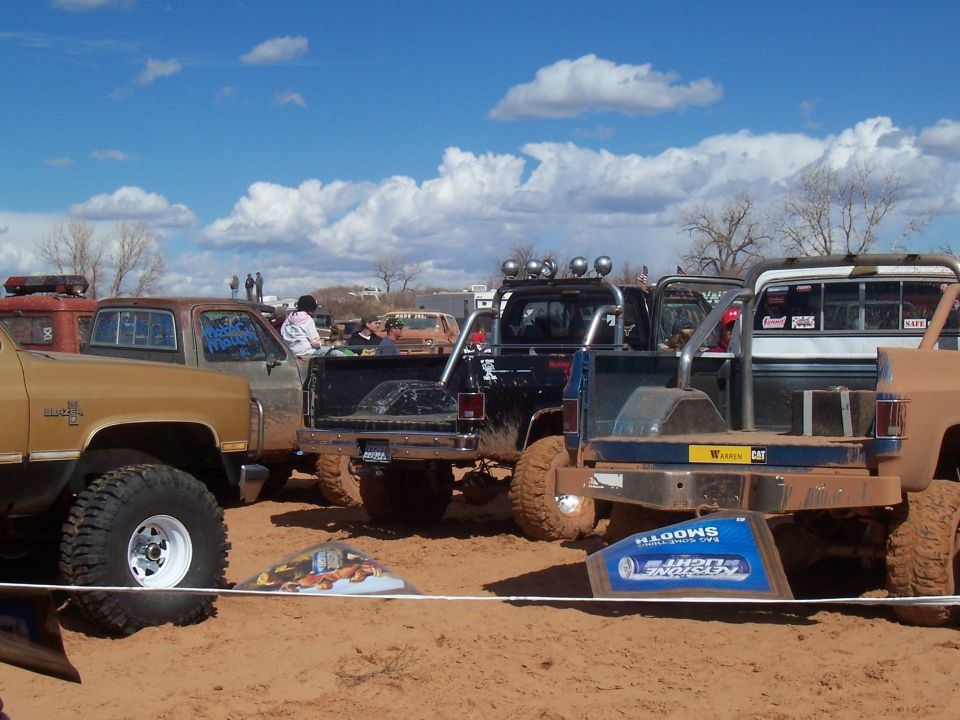 Several off-road pickup trucks parked in a sandy, muddy area under a cloudy sky. One truck has a Bud Light sign on its tailgate