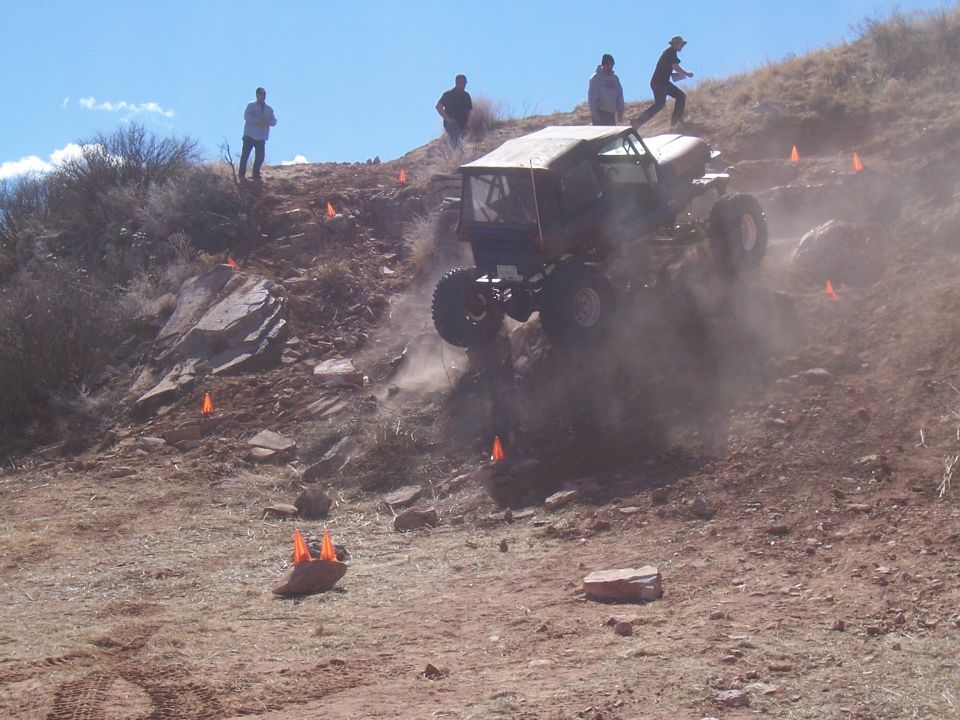 Off-road vehicle with large tires climbing a dusty, rocky hill with spectators watching