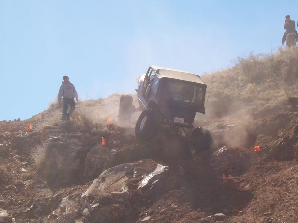Off-road vehicle with a soft top navigating a steep, dusty hill with orange markers