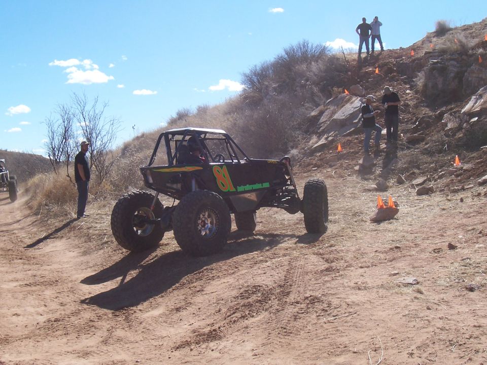 Off-road buggy with number 81 and website text on the side, parked on a dirt track with people and orange cones on a rocky hill