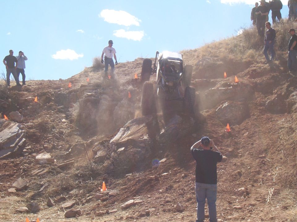 Off-road buggy climbing a steep, rocky hill with orange cones marking the course. Spectators watch from the side