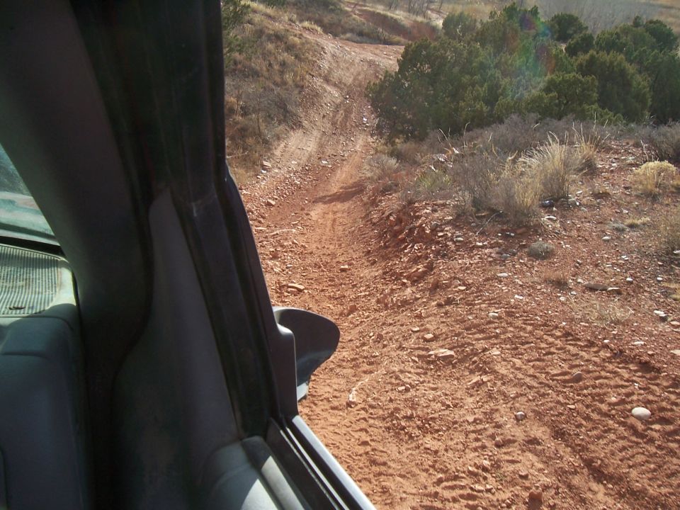 View from inside a vehicle looking out at a dusty, rutted dirt road winding through a dry, scrubby landscape