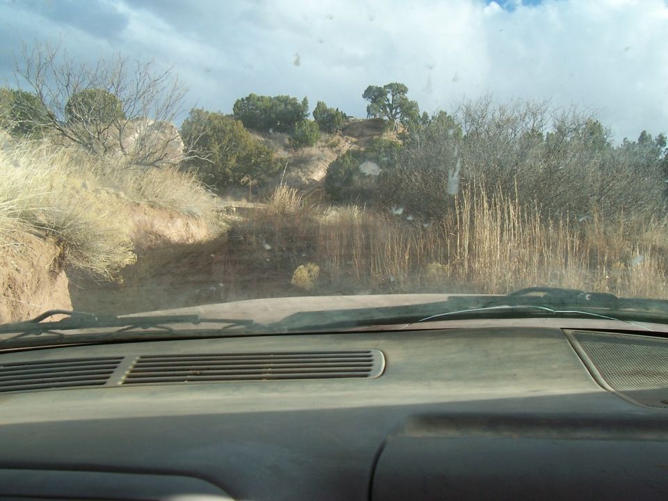 View from inside a vehicle looking out at a dusty, dry landscape with sparse vegetation and a cloudy sky