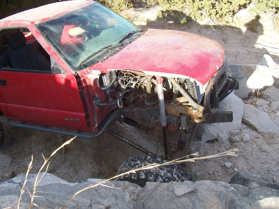 Red pickup truck with front end damage, missing bumper and hood, off-road tires, and side protection bars