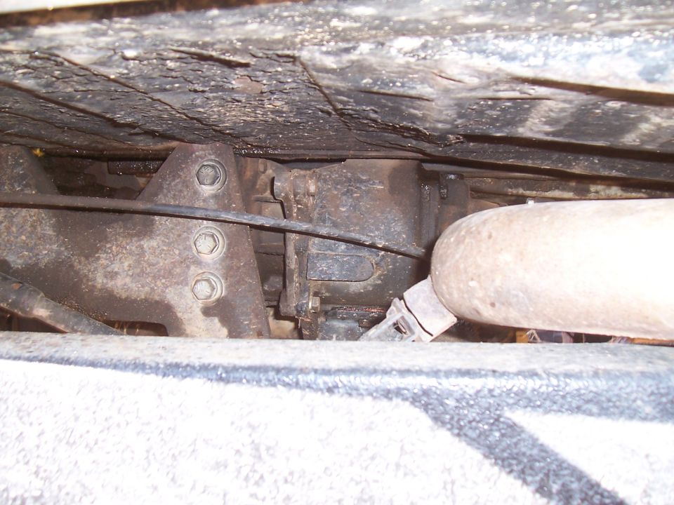 Underside view of vehicle components, including a rusty bracket, part of the drivetrain, and an exhaust pipe