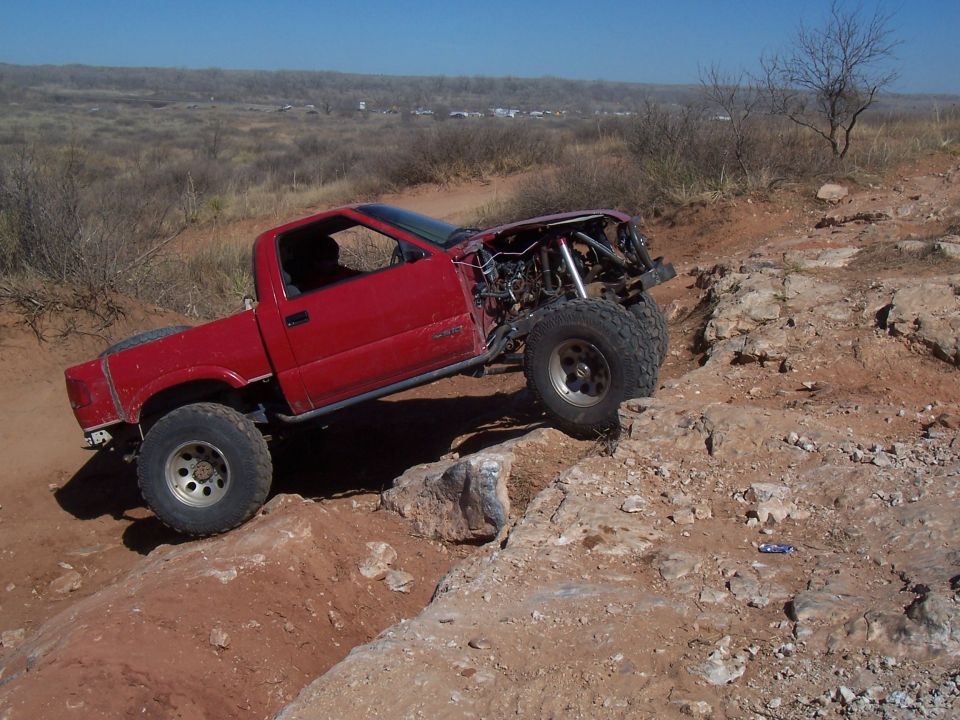 Red pickup truck with modified front end and large tires navigating a rocky trail