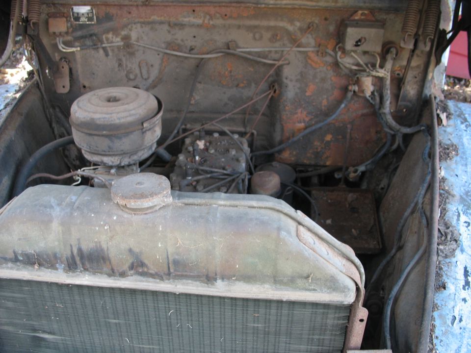 Dusty vintage engine bay with rusty radiator, round air cleaner, and inline engine