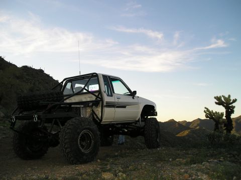 1986 Toyota Hilux - Modified 1986 Toyota Hilux with a roll cage and oversized tires sits on a rocky incline under a cloudy sky.