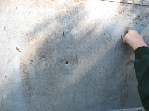 1952 Ford Panel Van - A close-up view of a damaged metal panel, possibly from a vehicle, with a bullet hole and a hand interacting with the surface.