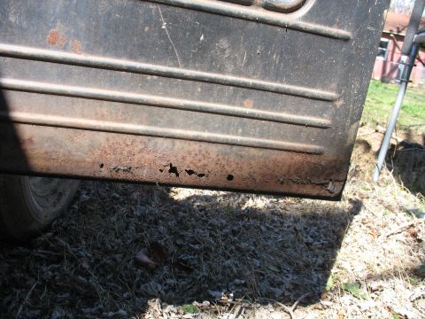 1952 Ford Panel Van - Close-up of the lower rear body panel of an old dark-painted vehicle showing heavy rust perforation and pitting along the bottom edge, with…