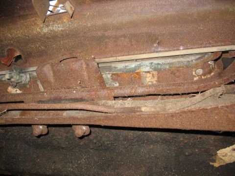 1952 Ford Panel Van - Close-up of a vehicle's heavily rusted underside, showing a corroded frame rail, a leaf spring, and a rusty body-mount bracket caked in dir…