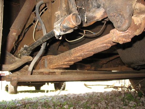 1952 Ford Panel Van - Underbody shot of a heavily rusted rear leaf-spring pack, axle housing and shackle on an old truck parked on leaves and grass. A black rubb…