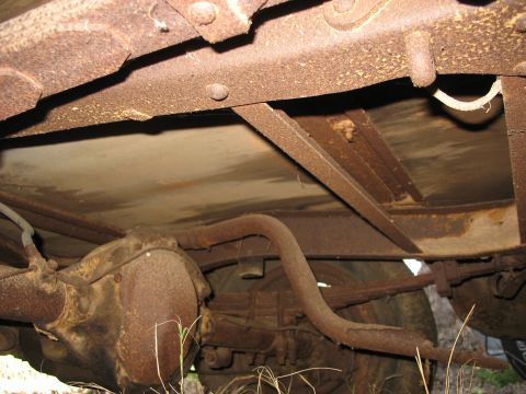 1952 Ford Panel Van - Underside view of a heavily rusted vintage vehicle chassis, showing the riveted ladder frame rail and crossmember, floor pan, leaf-spring r…