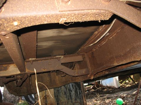 1952 Ford Panel Van - Underside view of a heavily rusted vintage vehicle body resting on wood blocks, showing the boxed frame rail, riveted crossmember and rotte…