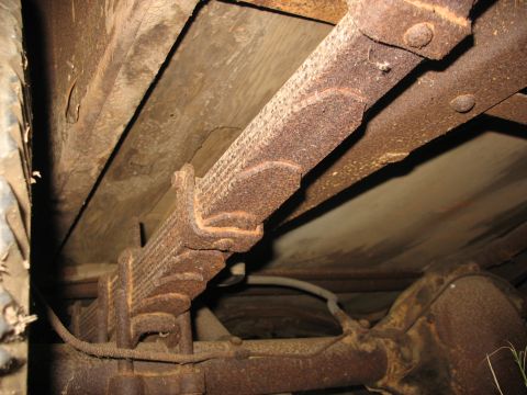 1952 Ford Panel Van - Underbody shot of a heavily surface-rusted leaf spring pack and shackle mount running along the frame rail, with the wooden bed floor plank…