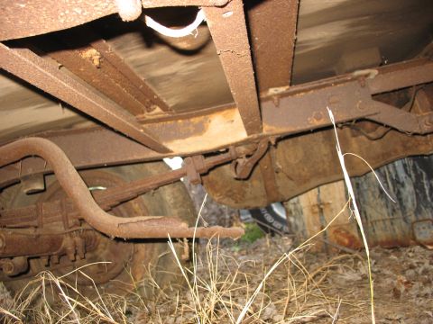 1952 Ford Panel Van - Underside view of an old truck project sitting in dry grass, showing a heavily rusted frame rail, floor crossmembers, leaf spring, rear axl…