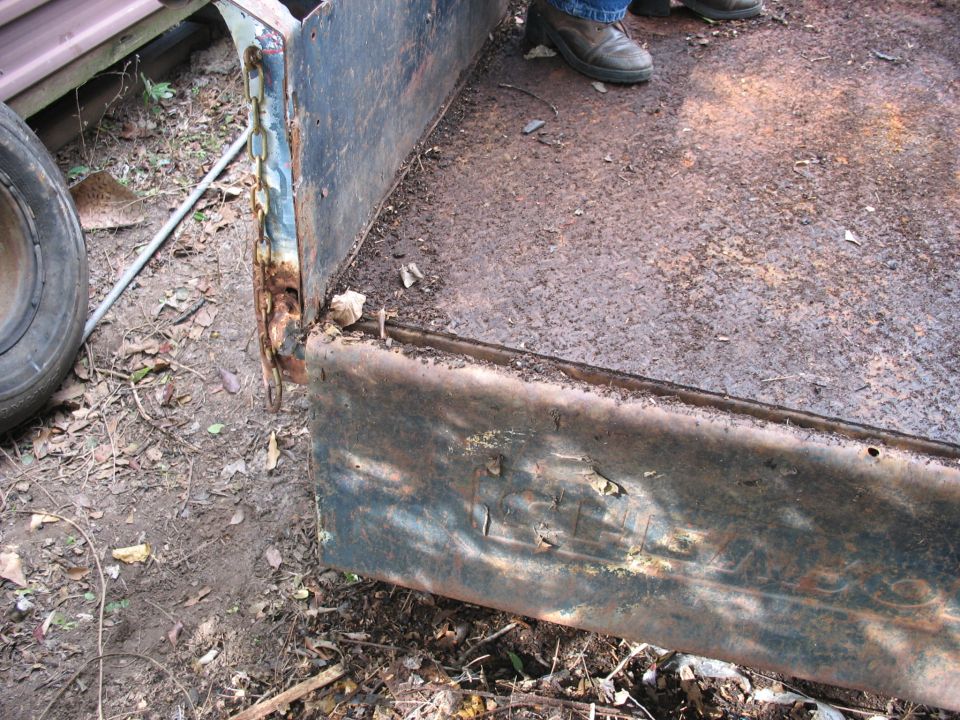 Close-up of a rusty truck bed with a chain hanging off the side. A person's legs and boots are visible standing in the bed