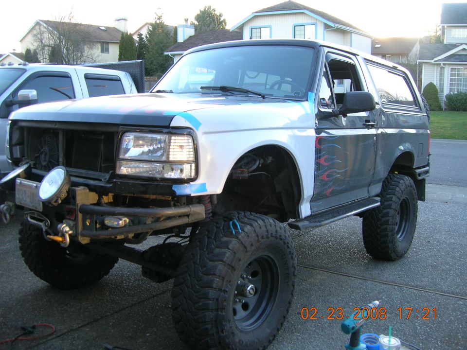 Lifted white Ford Bronco with flame graphics, grille removed, winch bumper and big mud tires