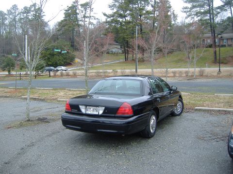 2001 Ford Crown Victoria - A black 2001 Ford Crown Victoria is parked in a gravel lot with trees and houses in the background.