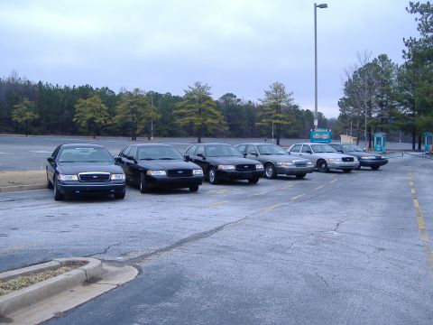 2001 Ford Crown Victoria - A line of black Ford Crown Victorias are parked in an outdoor lot under an overcast sky.