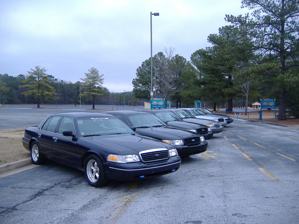 Several dark Ford Crown Victorias parked in a lot. The front one has aftermarket wheels