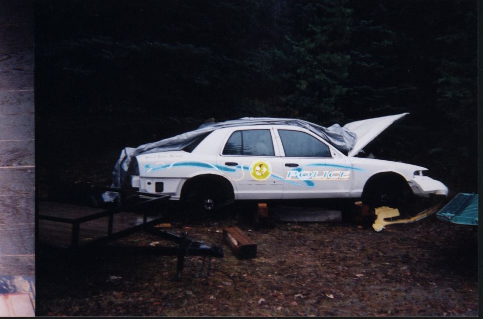 White Crown Victoria police sedan on blocks with wheels removed, hood open, tarp on rear, in wooded yard