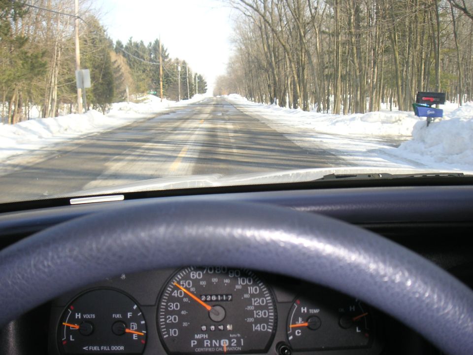 View from inside a car, looking out at a snowy road ahead. Steering wheel and dashboard visible