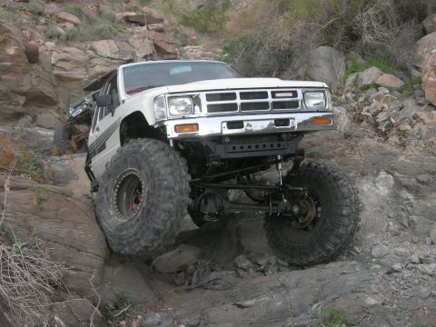 1986 Toyota Hilux - Modified white Toyota pickup truck navigates rocky terrain, showing its lifted suspension and oversized tires.