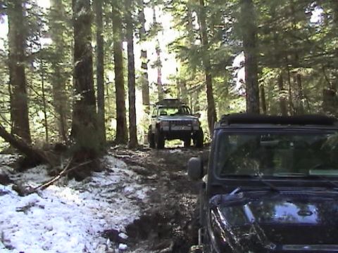 2005 Jeep Wrangler - A black soft-top Jeep Wrangler sits at right on a muddy, patchy-snow forest trail while a white Jeep Cherokee XJ with roof rack faces it fr…