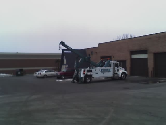 White tow truck with a green boom, parked in front of a brick building