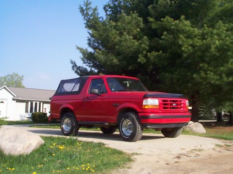 1995 Ford Bronco - A red Ford Bronco with a removable soft top is parked on a dirt road in front of a house and trees.