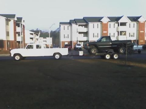 1978 Ford Bronco - White crew cab pickup truck is towing a dark pickup truck on a trailer.