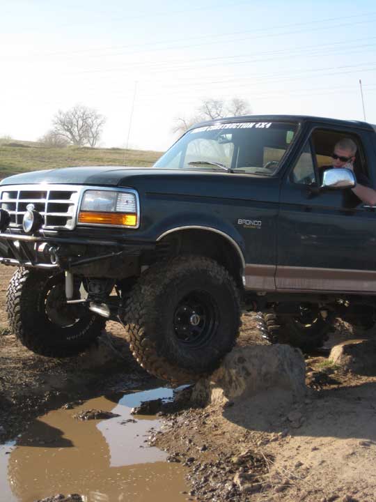 Dark green Ford Bronco with lifted suspension navigating muddy terrain