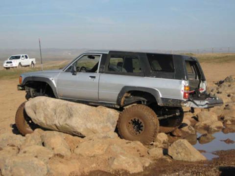 1978 Ford Bronco — Silver Toyota 4Runner on rocks with a white pickup truck in the background