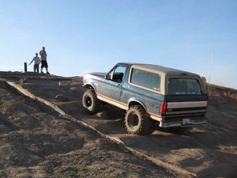 1978 Ford Bronco — Blue and tan SUV with large tires driving on a dirt hill