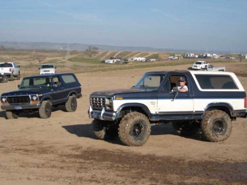 Two Ford Broncos in a dirt lot, one blue and white, the other dark blue. The blue and white Bronco is in the…