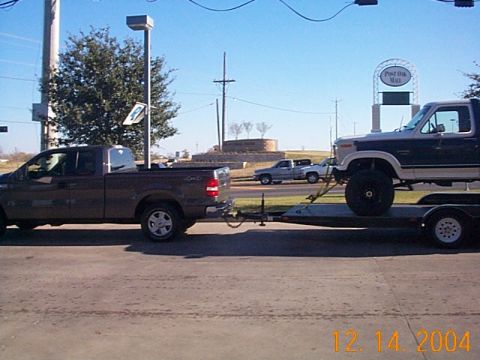 2004 Ford F-150 - A brown pickup truck tows a lifted Ford F-250 Super Duty on a trailer down a road.
