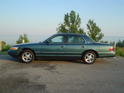 1996 Mercury Grand Marquis - A green sedan is parked on a road near a body of water under a clear sky.