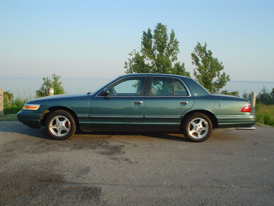 Green sedan parked on asphalt with water and trees in the background