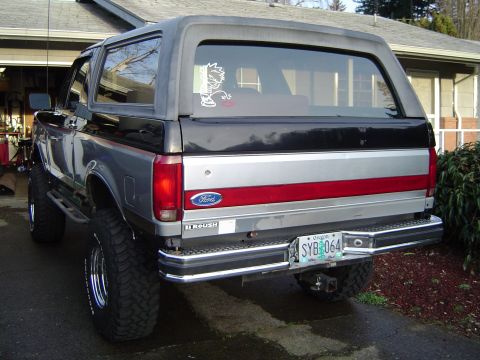 1989 Ford Bronco - The rear of a two-tone Ford Bronco is parked in a driveway, showing its license plate and a humorous sticker.
