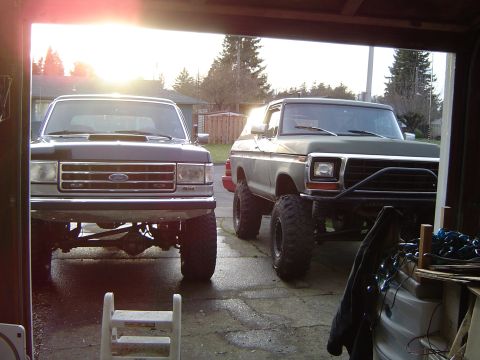 1989 Ford Bronco - Two Ford Broncos, one dark grey and one olive green, are parked side-by-side in a garage.