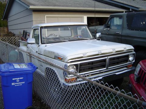 1989 Ford Bronco - A white Ford pickup truck is parked in a driveway next to a garage, partially obscured by a chain-link fence.