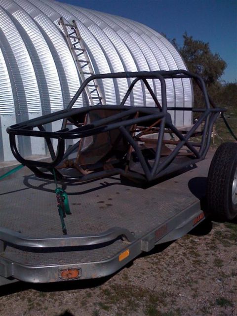 1994 Ford Bronco - The frame of an off-road buggy is loaded onto a trailer, with a large corrugated metal building in the background.