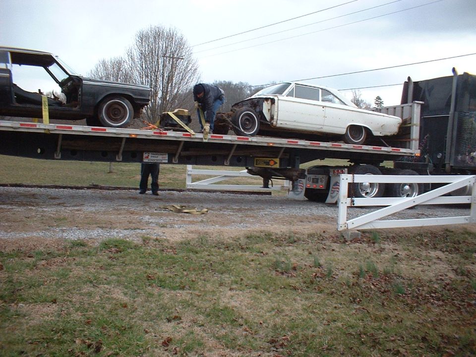 Two old cars, one black and one white, being transported on a flatbed trailer. A person is securing the white car